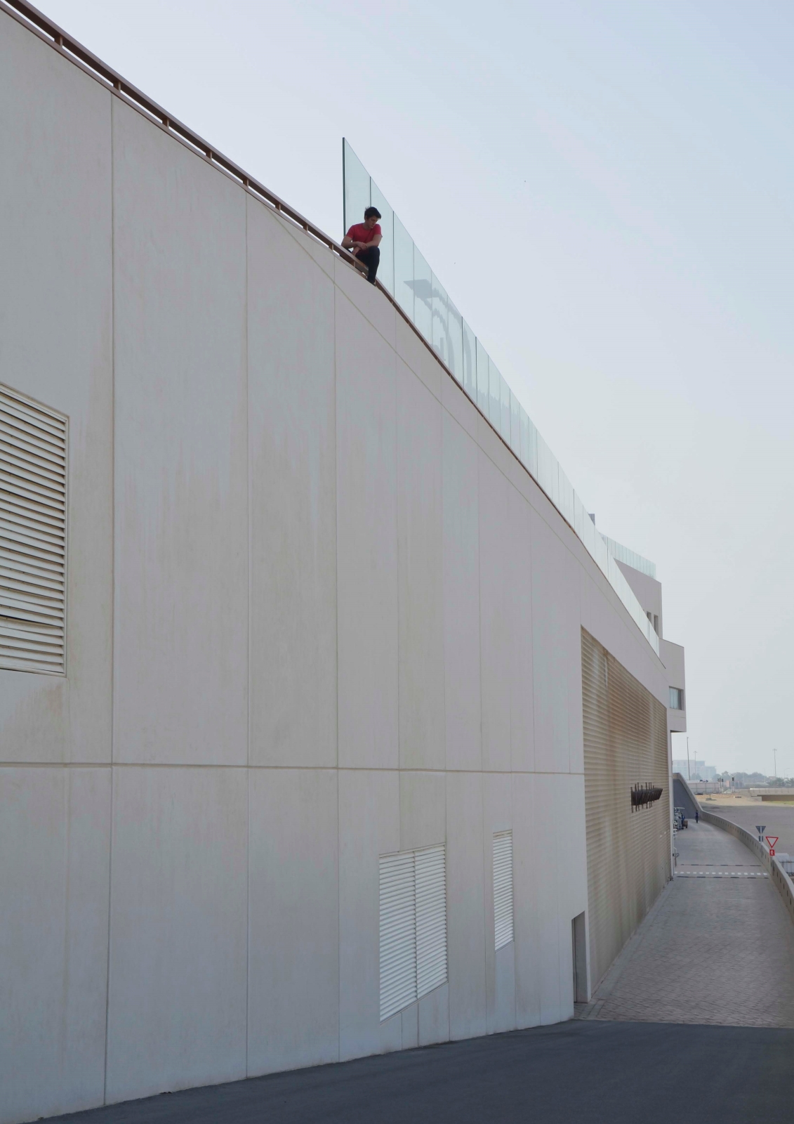 A man in a pink shirt sits atop a high wall above a road.