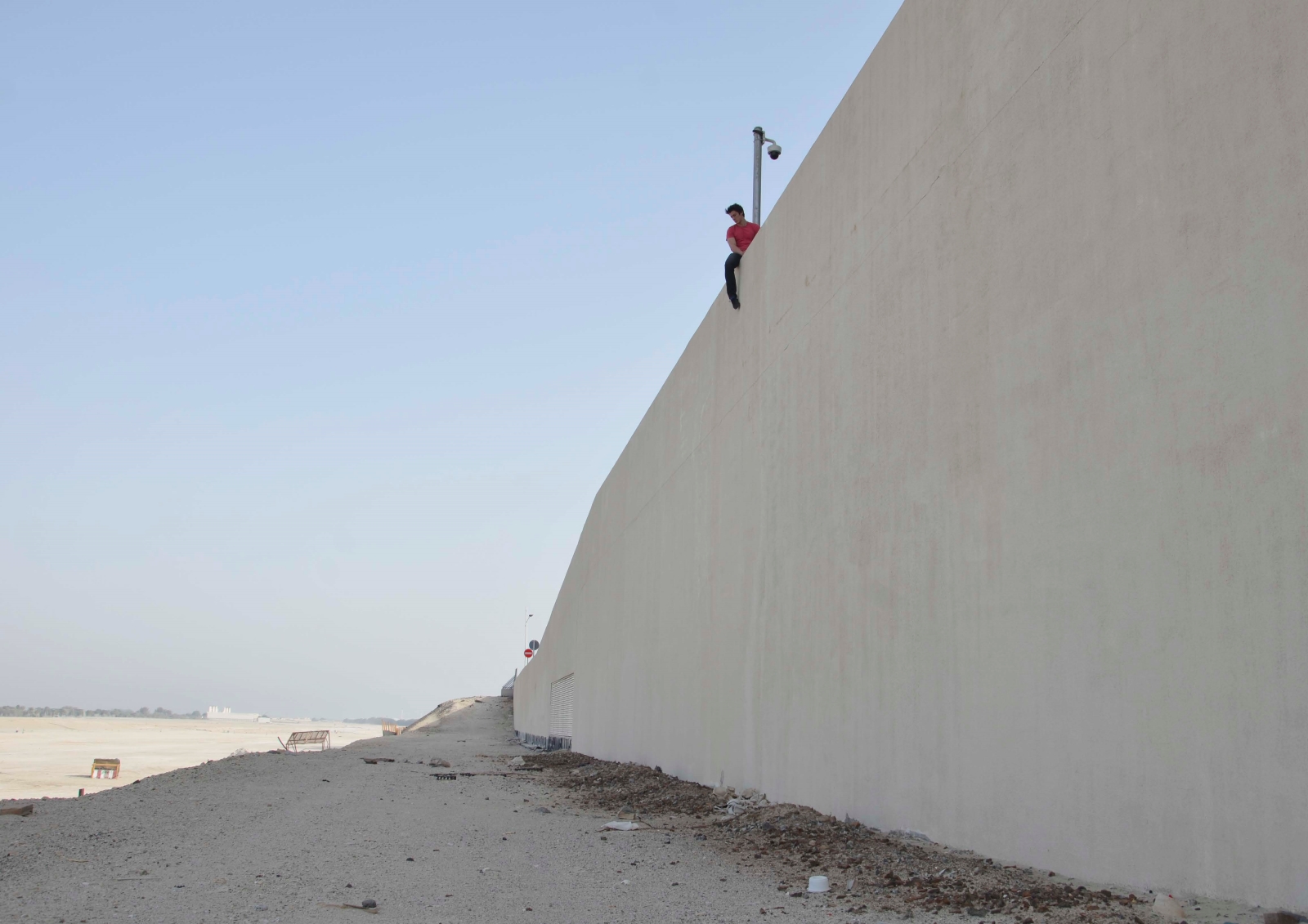 A man in a pink shirt sits atop a high wall above a desert.