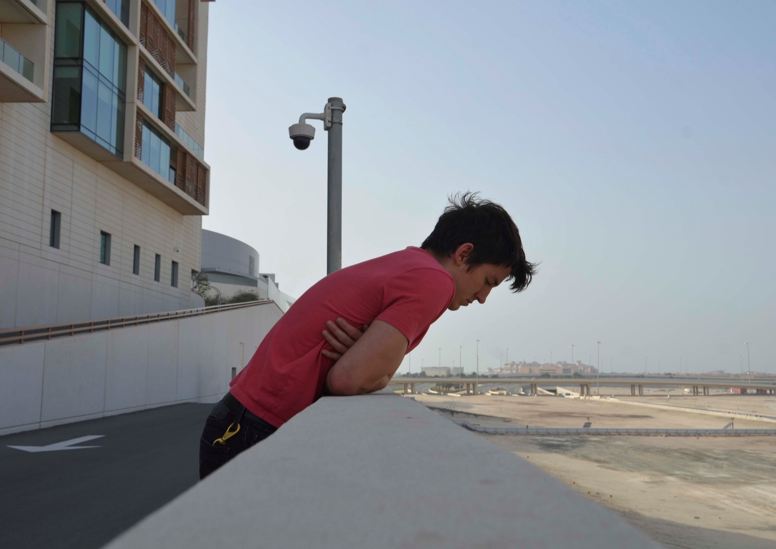 A man in a pink shirt leans over the edge of a wall. A surveillance camera can be seen behind him.